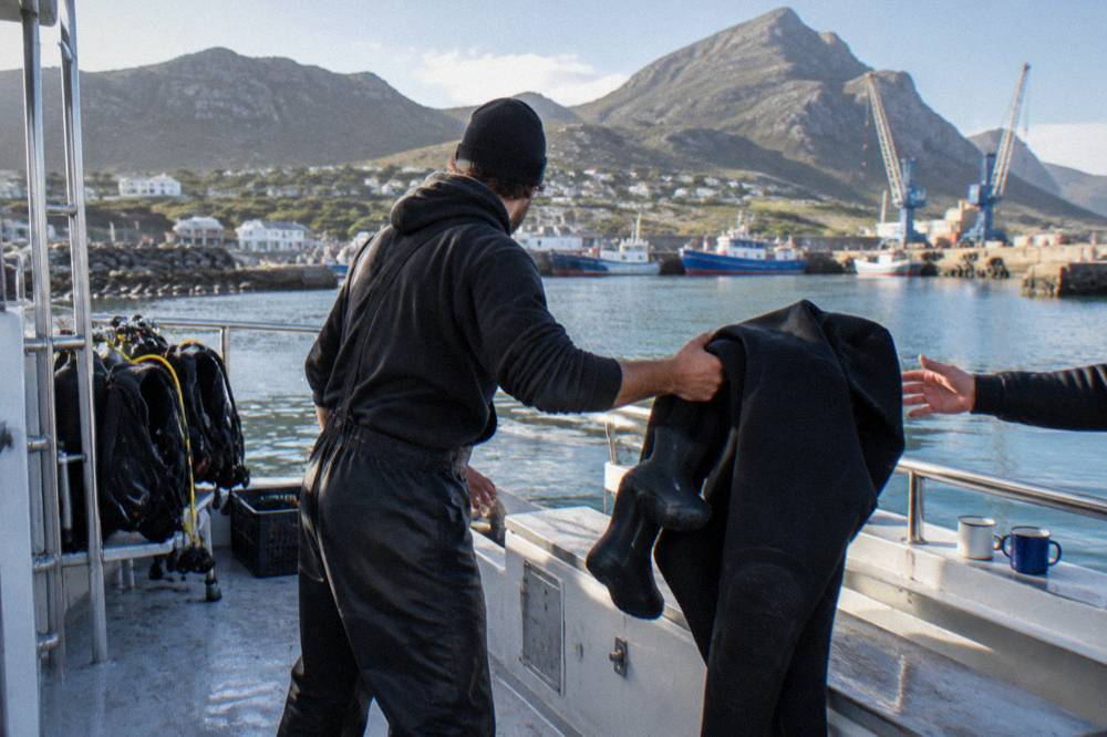 A dive crew member handing a thick black wetsuit and booties across the deck of a shark diving boat