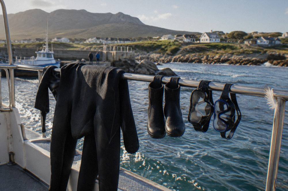 A dark neoprene wetsuit hanging to dry on a railing next to dive boots and masks