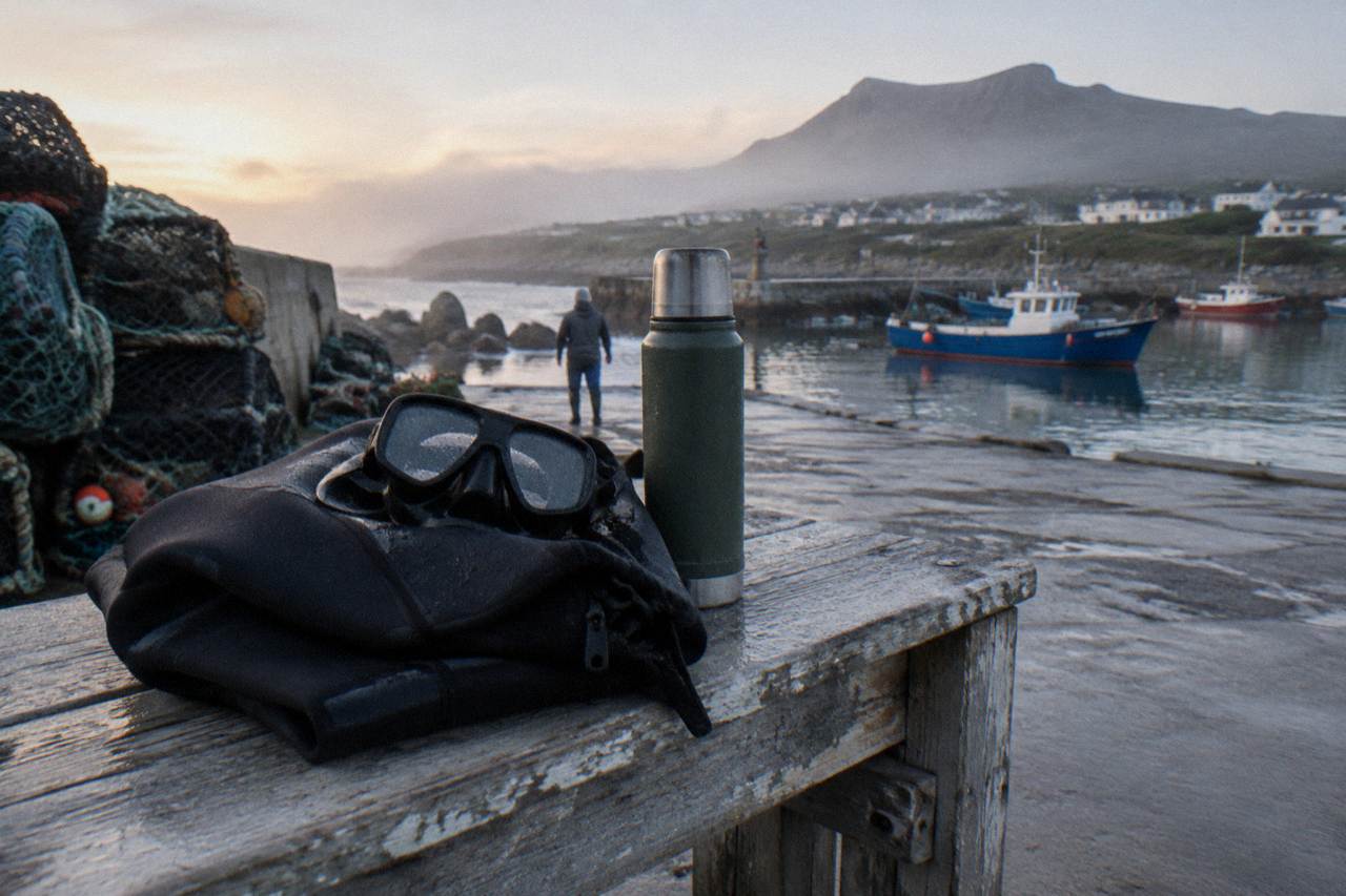 A folded black wetsuit and dive mask resting on a wooden bench beside a harbour deck at dawn