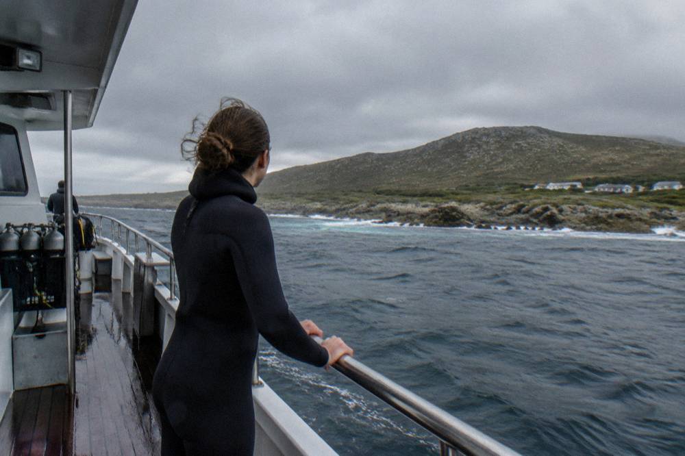 A passenger in a wetsuit holding the rail of a boat while looking toward the distant coastline