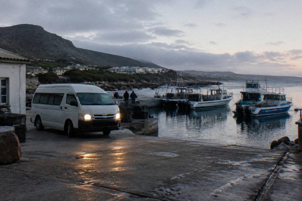 A tour minibus parked at Kleinbaai Harbour with shark cage diving boats in the background