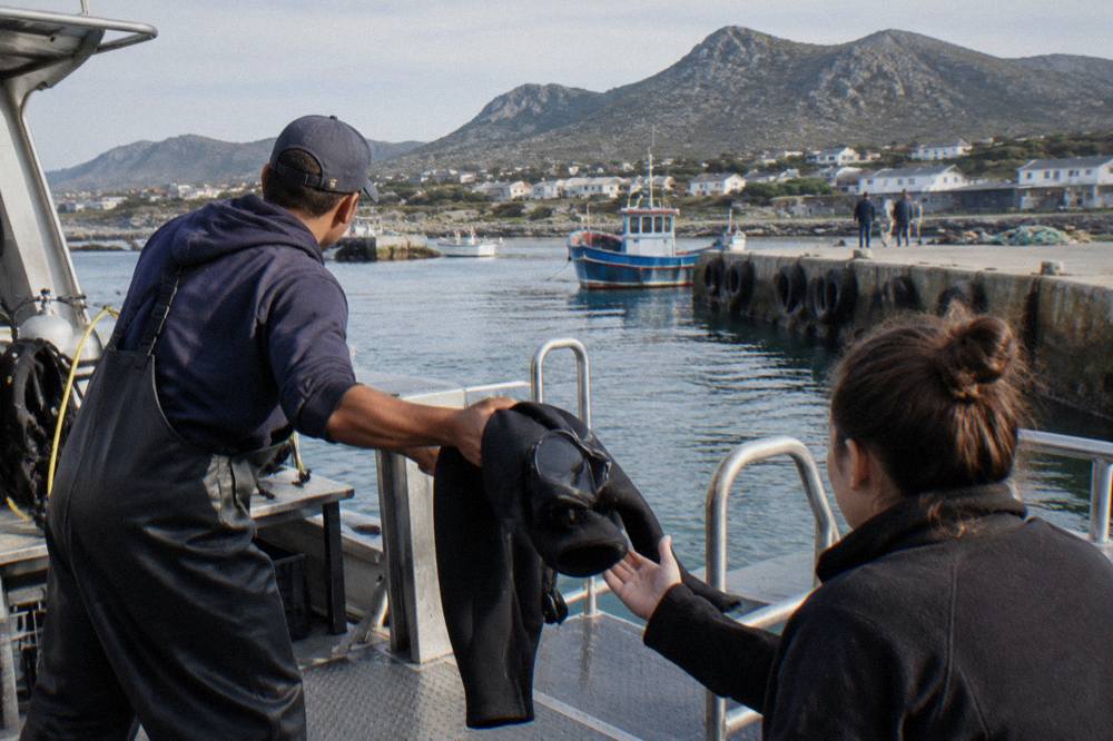 A dive crew member handing a wetsuit and mask to a passenger on the deck of a shark diving boat